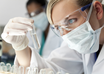 A blond medical or scientific researcher or doctor using looking at a clear solution in a laboratory with her Asian female colleague out of focus behind her.