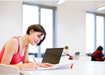 Pretty, young college student studying in the library/a study room at campus , using her modern laptop computer, writing notes, preparing for exams (shallow DOF)