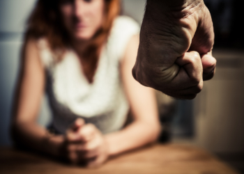 Young woman is sitting hunched at a table at home, the focus is on a man's fist in the foregound of the image